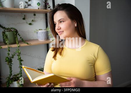 Nachdenkliche Frau, die ein offenes Notizbuch neben Zimmerpflanzen zu Hause hält. Offline-Studium, reflektierendes Notizenmachen und persönliches Wachstum. Stockfoto