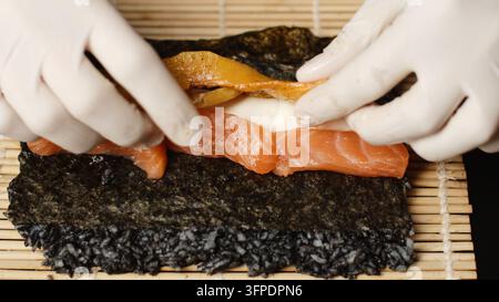 Chef gibt Handschuhe an, bereitet frische Lachssushirolle mit schwarzem Reis zu, fügt Zutaten auf Nori und Bambusmatte hinzu Stockfoto