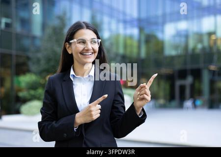 Junge Geschäftsfrau in Anzug und Brille, die glücklich lächelt und mit den Fingern zur Seite zeigt, selbstbewusst vor einem modernen Firmengebäude stehend Stockfoto