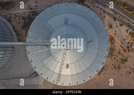 Luftaufnahme eines großen runden Metallgetreidesilos mit einem zentralen Förderband und einer Leiter, umgeben von trockenem Boden und Bahngleisen in Eaton, Col Stockfoto