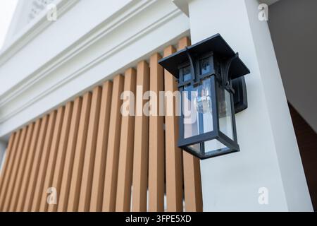 Schwarze Wandlaterne im Vintage-Stil mit Glasplatten und Glühbirne mit Filament, montiert auf weißem Pfeiler eines modernen Hauses, Holzlatten dahinter, Außenbeleuchtung Stockfoto