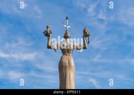 Die Statue einer Frau, die zwei kleine Figuren hochhält, von hinten aufgenommen vor einem blauen Himmel, Imperia-Statue im Hafen von Konstanz am Bodensee, B Stockfoto