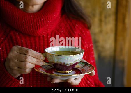 Frau in rotem Strickpullover bietet eine schöne Vintage-Porzellan-Teetasse voller heißem Tee an Stockfoto