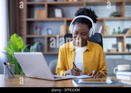 Lächelnde Frau trägt Kopfhörer und benutzt einen Laptop für Fernunterricht, Telearbeit oder die Teilnahme an einem Webinar, schreibt in ein Notizbuch an einem Schreibtisch im Homeoffice Stockfoto