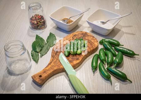 Frische grüne Jalapeno-Pfeffer und in Scheiben geschnittene Jalapenos, die für die Konservierung in Salzlake vorbereitet wurden, mit Lorbeerblättern, Fenchelsamen, gemischten Pfefferkörnern, Salz und Glasvorratsgläsern, die auf einem Küchentisch angeordnet sind Stockfoto