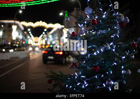 Eine festliche Abendszene zeigt eine mit lebhaften Weihnachtslichtern geschmückte Straße. Ein Weihnachtsbaum glitzert mit Schmuck. Autos fahren vorbei, während Menschen en Stockfoto