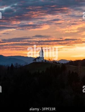 Luftaufnahme einer Kirche, die auf einem grasbewachsenen Hügel bei Sonnenuntergang mit einem dramatischen orangefarbenen Himmel und fernen Bergen in Slowenien thront Stockfoto