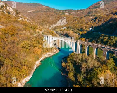 Luftbild der Solkan-Brücke, die den smaragdgrünen Soca-Fluss überspannt, umgeben von herbstfarbenen Wäldern und steilen Hügeln in Slowenien. Stockfoto