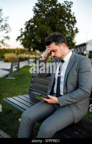 Ein Mann in einem Anzug sitzt auf einer Bank in einem Park. Er sieht gestresst aus, während er sein Tablet benutzt. Stockfoto