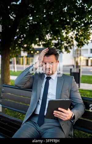Ein Mann im Anzug sitzt tagsüber auf einer Bank in einem Park. Er sieht besorgt aus, während er ein Tablet benutzt. Stockfoto