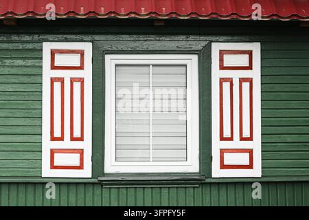 Weißes Fenster mit roten dekorativen Fensterläden an einer dunkelgrünen Holzfassade. Architektur-Hintergrund für traditionelles Außendesign eines rustikalen Hauses. Stockfoto