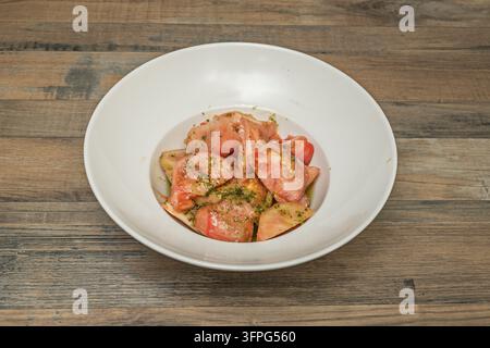 Frische Tomatenscheiben, beträufelt mit Olivenöl, Essig und gehackten Kräutern, serviert in einer breiten weißen Schüssel auf einem rustikalen verwitterten Holztisch Stockfoto