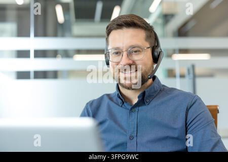Lächelnder Mann mit Headset und Mikrofon an einem Laptop, der an einem virtuellen Meeting teilnimmt, aus einem modernen Büro, selbstbewusster Profi, der Remote-Kundensupport oder Klientensupport bietet Stockfoto
