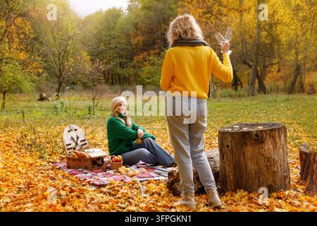 Gemütliches Herbstpicknick mit Freunden im goldenen Park: Frauen entspannen sich im Freien, halten Weingläser, stimmungsvolle Herbst-Fotosession im Wald Stockfoto