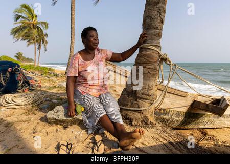 Eine Frau sitzt neben einem hölzernen Fischerboot unter einer Palme am Strand, Zentralregion, Elmina, Ghana Stockfoto