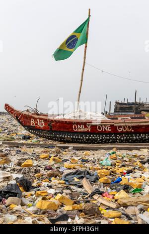 Ein bemaltes Holzfischerboot mit einer brasilianischen Flagge liegt an einem mit Plastikmüll bedeckten Strand in der Greater Accra Region, Accra, Ghana Stockfoto