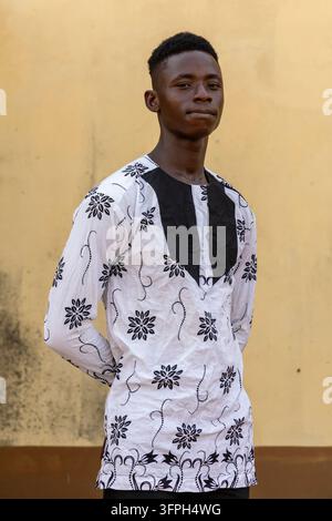 Porträt eines jungen Mannes, der ein weißes traditionelles Hemd mit schwarzem Blumenmuster vor einer gelben Wand trägt, Zentralregion, Elmina, Ghana Stockfoto