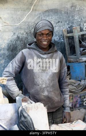 Lächelnder junger Arbeiter in einem staubigen grauen Hoodie und Kopftuch, der mit einer Hand in der Hüfte in einer Werkstatt im Kibera-Viertel steht und Souvenirs für Touristen herstellt aus Stockfoto
