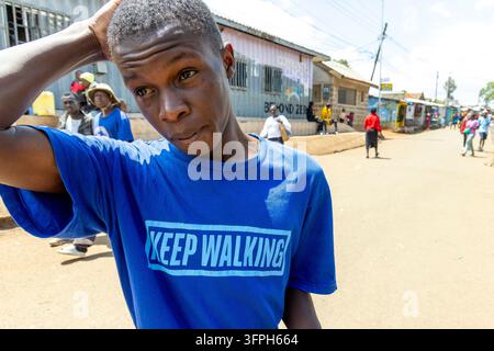 Junger Mann trägt ein Keep Walking T-Shirt mit einer Hand auf dem Kopf auf einer unbefestigten Straße im Kibera-Gebiet, Nairobi County, Nairobi, Kenia Stockfoto