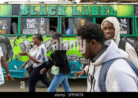 Junge Männer und Frauen gehen eine Straße entlang an einem grünen Matatu-Minibus mit Black Label-Schriftzug und Graffiti-Aufklebern vorbei, Nairobi County, Nairobi, Kenia Stockfoto