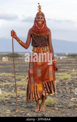 Lächelnde Frau des Turkana-Stammes mit roten Perlenketten, einem Kopfschmuck und einem beperlten Ledersaum, die mit einem langen Stock auf felsigem Boden mit Bergen steht Stockfoto