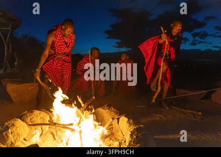 Vier Maasai-Männer um ein Lagerfeuer in der Dämmerung mit Stöcken und roten Shukas, Narok County, Maji Moto, Kenia Stockfoto