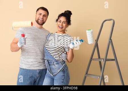 Hausrenovierung und DIY-Projekt, glückliches Paar hält Farbrollen mit Leiter für die Hausverbesserung und Innenarchitektur-Arbeiten auf einem beigen Hintergrund Stockfoto
