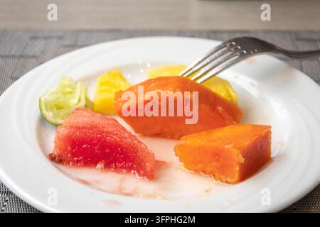 Frischer tropischer Obstteller mit Papaya, Wassermelone, Ananas und Limette auf einem weißen Teller, halb gegessen Stockfoto
