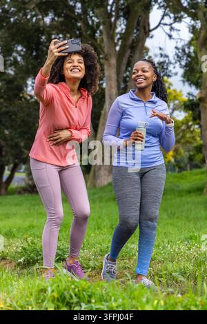 Verschiedene Freundinnen gehen im Park in Sportkleidung und machen mit dem Handy ein Selfie mit wiederverwendbarer Flasche. Draußen, Grünflächen, Bäume, Fitness, Hydration, Natur, lässig Stockfoto