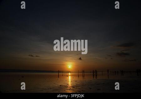 Goldener Sonnenuntergang über einem Strand in Bali, Indonesien, mit Menschen, die sich als Silhouetten vor dem Horizont abheben, und Sonnenlicht, das sich auf nassem Sand spiegelt Stockfoto