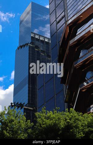 Die kupfernen Treppen des Vessel erheben sich neben reflektierenden Wolkenkratzern in Hudson Yards, eingerahmt von laubenden Bäumen und einem auffälligen blauen Manhattan-Himmel. Stockfoto