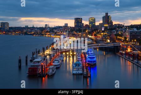 Blaue-Stunden-Blick über den Hafen von Hamburg mit beleuchteten Landungsbrücken, Passagierbooten und der Stadtsilhouette, die sich in der Elbe spiegelt. Stockfoto