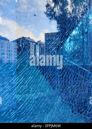 Eine Nahaufnahme eines schwer beschädigten und zertrümmerten blauen Glasfensters, das komplexe Bruchmuster zeigt, die die Reflexionen der städtischen Bebauung verzerren Stockfoto
