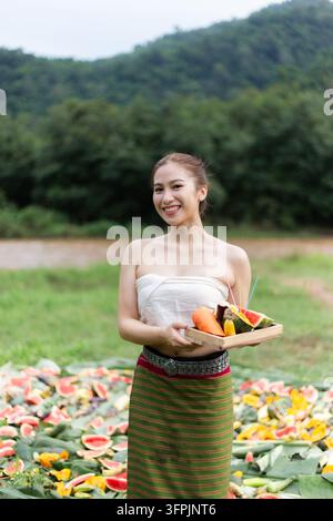 Eine junge asiatische Frau hält ein Obsttablett im Freien mit Bergen und Wassermelonenausbreitung im Hintergrund Stockfoto