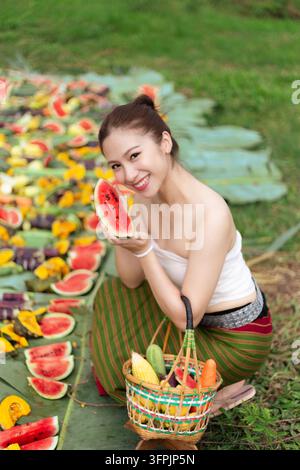 Eine junge asiatische Frau hält eine Wassermelone neben einem Gemüsekorb im Garten und lächelt während der Ernte im Freien Stockfoto