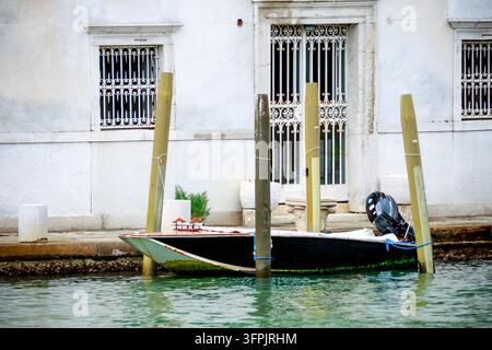 Kleines Holzboot, das an Kanalpfeilern neben einem verwitterten weißen Gebäude mit vergitterten Fenstern in Venedig, Italien, festgemacht ist. Stockfoto