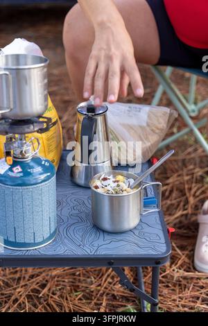 Person, die nach einer Moka-Kanne auf einem Campingtisch greift und ein gesundes Frühstück im Freien genießt. Freiheit und Abenteuer erleben Stockfoto