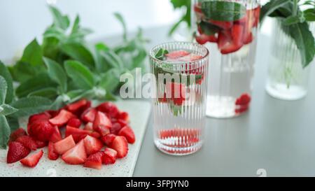 Frisches Erdbeer- und Minz-Detoxwasser mit Eiswürfeln, serviert in Gläsern auf dem Küchentisch, umgeben von geschnittenen Beeren und Minzblättern. Stockfoto