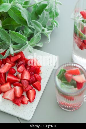 Frisches Erdbeer- und Minz-Detoxwasser mit Eiswürfeln, serviert in Gläsern auf dem Küchentisch, umgeben von geschnittenen Beeren und Minzblättern. Stockfoto