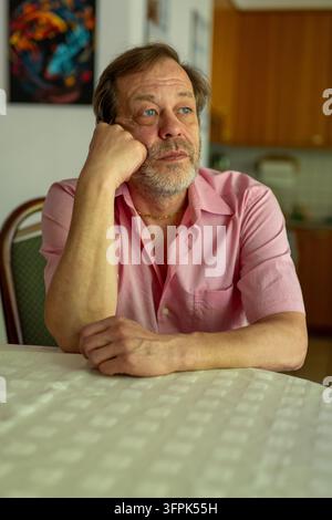 Ein mittelalter Mann in einem rosa Hemd stützt seinen Kopf auf die Hand an einem Tisch im Innenraum. Stockfoto