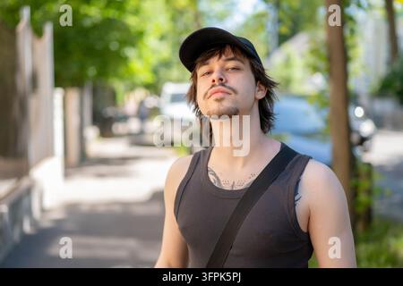 Ein junger Mann mit einer schwarzen Kappe und einem Tanktop posiert an einem sonnigen Stadtrandstein. Stockfoto