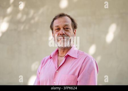 Ein lächelnder Mann mittleren Alters in einem rosa Hemd steht vor einer sanft verschwommenen Wand. Stockfoto