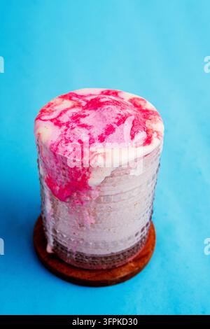 Hochwinkelaufnahme eines Erdbeersmoothie-Milkshakes im Glas mit übergelaufenem Schaum auf einem Holzuntersetzer vor blauem Hintergrund. Stockfoto