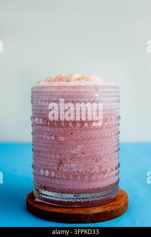 Hochwinkelaufnahme eines Erdbeersmoothie-Milkshakes im Glas mit übergelaufenem Schaum auf einem Holzuntersetzer vor blauem Hintergrund. Stockfoto