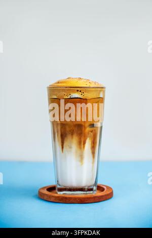 Eislatte-Kaffee in einem hohen Glas mit geschichteter Milch und Espresso, oben mit dickem Schaum, auf einem Holzuntersetzer über blauer Oberfläche. Stockfoto