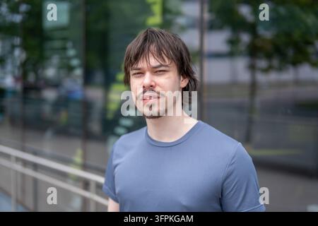Ein junger Mann mit mittellangem Haar und einem Kinnbart steht im Freien neben einem Glasgebäude. Stockfoto