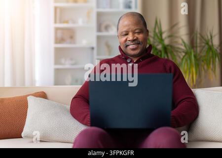Lächelnder schwarzer Senior entspannt sich auf dem Sofa zu Hause und arbeitet am Laptop Stockfoto