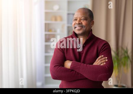 Selbstbewusster schwarzer Senior steht zu Hause mit verschränkten Armen und schaut weg Stockfoto