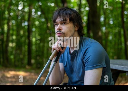Ein junger Mann sitzt im Wald im Freien, hält Trekkingstöcke und schaut nachdenklich weg. Stockfoto