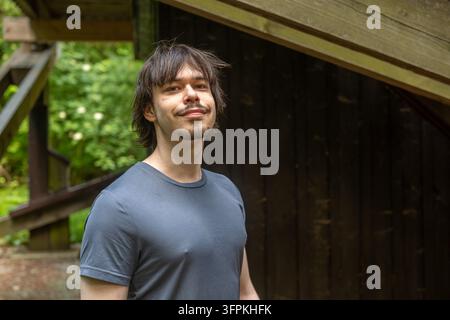 Ein lächelnder junger Mann steht im Freien neben einem dunklen Holzunterstand in einer grünen Waldkulisse. Stockfoto